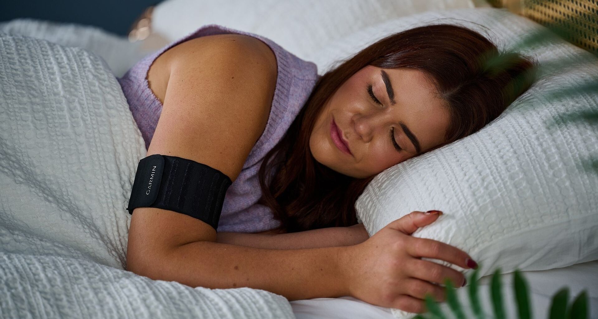 Our sleep tech reviewer laying in bed with white sheets wearing purple jumper and the black Garmin Index Sleep Monitor on right arm