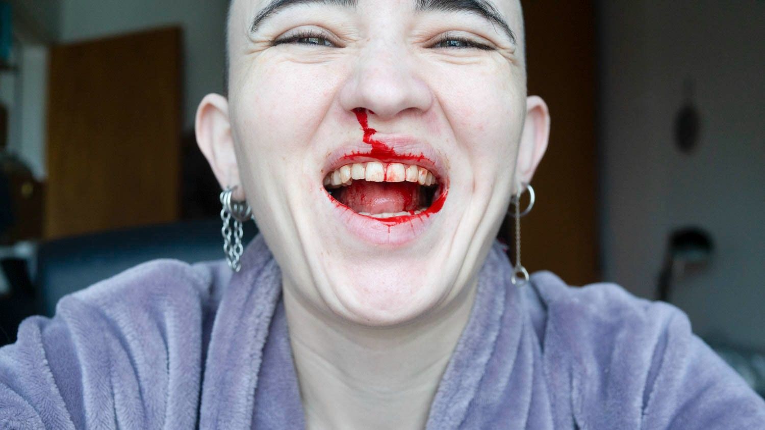 A close-up, joyful portrait of a person with a shaved head laughing broadly, with red liquid resembling blood smeared across their nose, lips, and teeth.