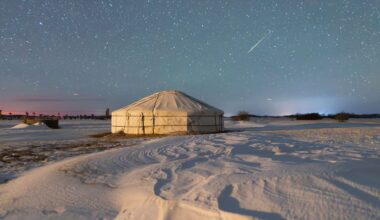 A meteor streaks throught the starry night sky above a fabric structure surrounded by snow.