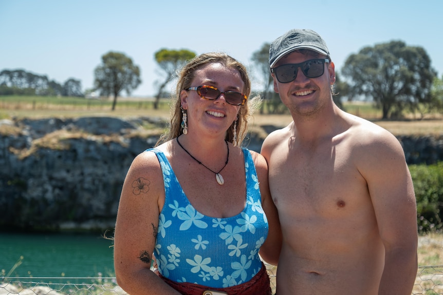 a woman and a man in swimwear and glasses smiling, with cliffs and a lake behind them