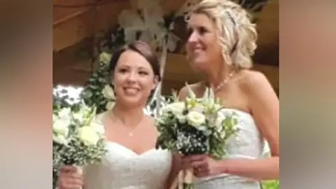 Family photo Helen and Rachael at their wedding wearing white strapless dresses and holding bouquets of white flowers