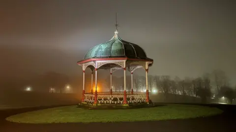 Fiona Mathieson A Victorian-style bandstand stands illuminated in a misty park at night. 