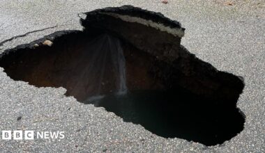 A residential street is cordoned off with blue-and-white police tape. A large hole or sinkhole is visible in the middle of the road.