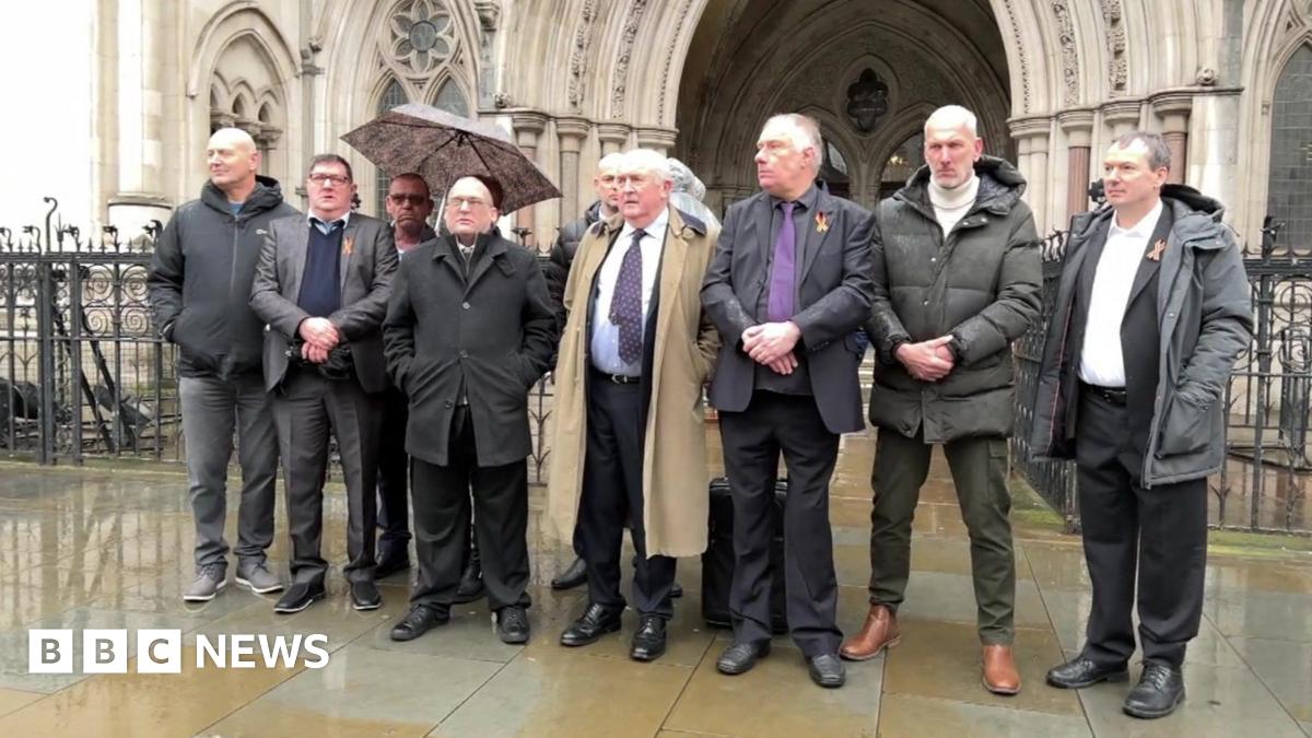 A group of men in smart suits and coats stand outside the entrance of the High Court, a grand, gothic building. They look solemn, and it appears to be raining.