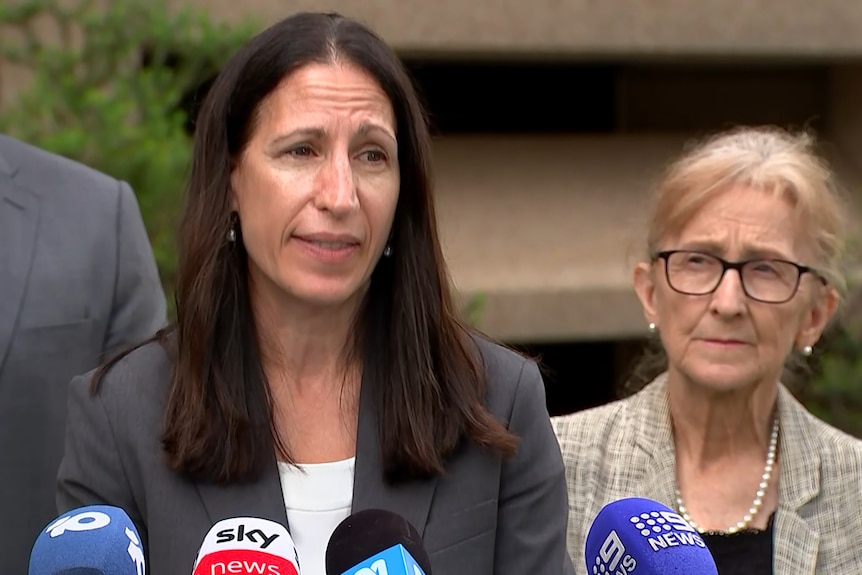 Michele Goldman front and centre at a press conference in a suit jacket and white top.