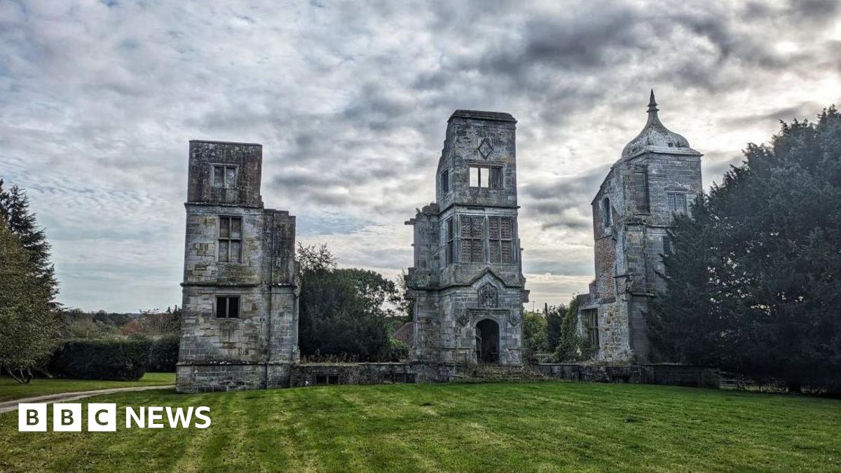 The image shows Brambletye House, three grey tower structures surrounded by trees and grass lawns. The structure is framed against a cloudy grey sky.