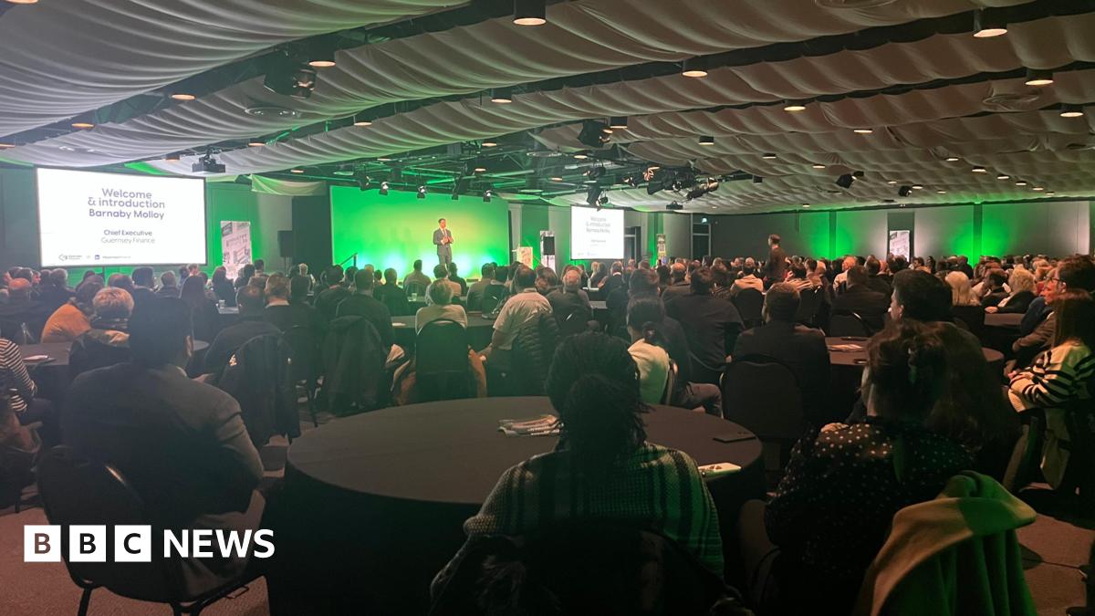 A man standing on a stage with a green background, with a number of tables well attended by people watching the speech.