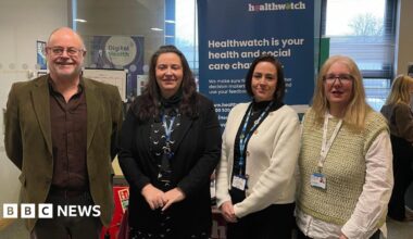 One man and three women stand in a line. They are the health hub team. They are standing in front of a Healthwatch banner.