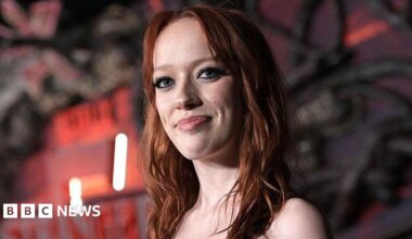 Marybeth is pictured at a film premier. There is a black and red background with strobe lighting. She is smiling at the camera and has ginger hair.