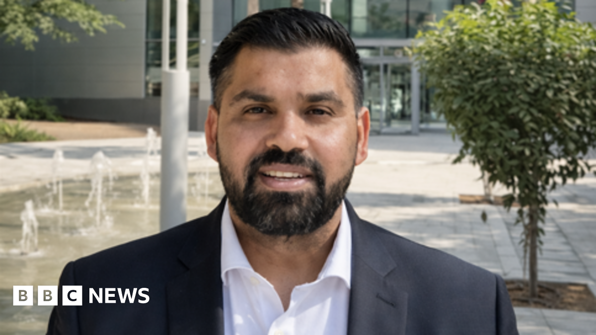 Mann is stood outside a building with a water fountain and small tree behind him. He has a navy suit on and a white shirt. He has dark hair and a beard.