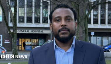 Doctor Awilly Chofle smiling at the camera. He is stood in front of Jersey General Hospital and is wearing a blue suit jacket with a light blue shirt.