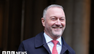 Secretary of State for Housing, Communities and Local Government Steve Reed walks across Downing Street. He is grinning, with closely cropped grey hair and stubble. He wears a navy overcoat, a white shirt and a red paisley tie.