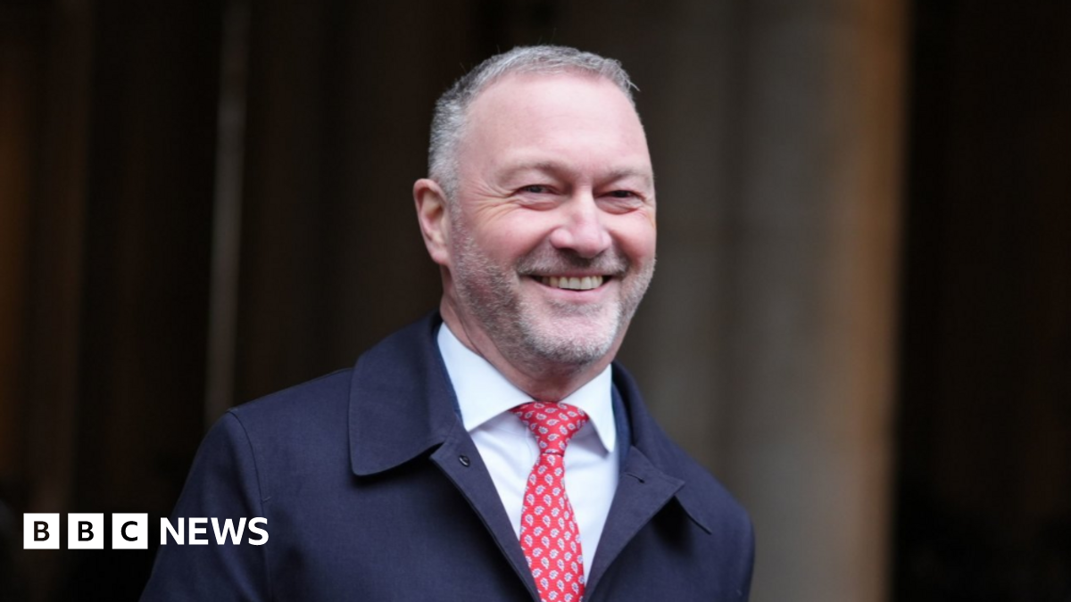 Secretary of State for Housing, Communities and Local Government Steve Reed walks across Downing Street. He is grinning, with closely cropped grey hair and stubble. He wears a navy overcoat, a white shirt and a red paisley tie.