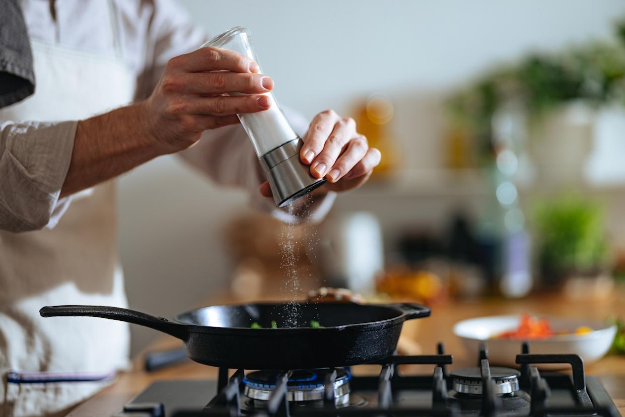 An anonymous chef with apron pouring salt into pan while standing at a gas stove.