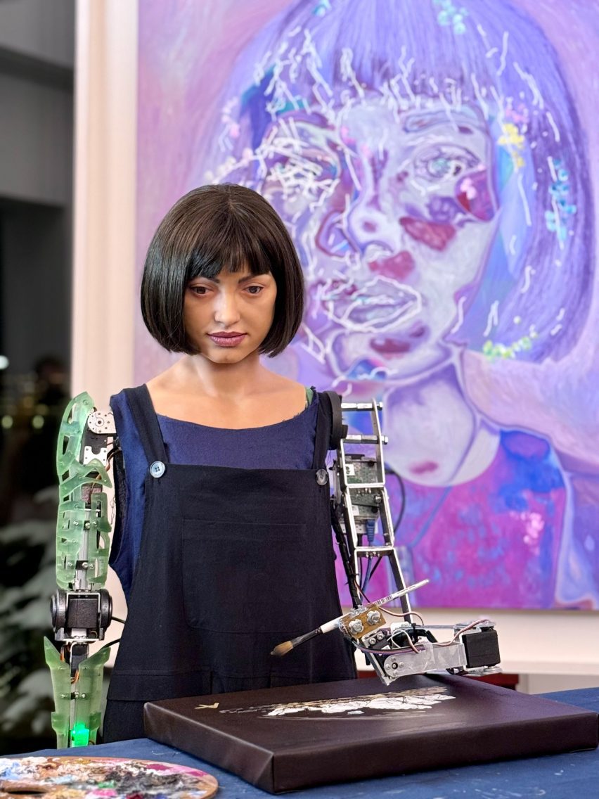 Photo of the Ai-Da humanoid robot with a paintbrush in her mechanised arm and an artwork in front of her and on the wall behind