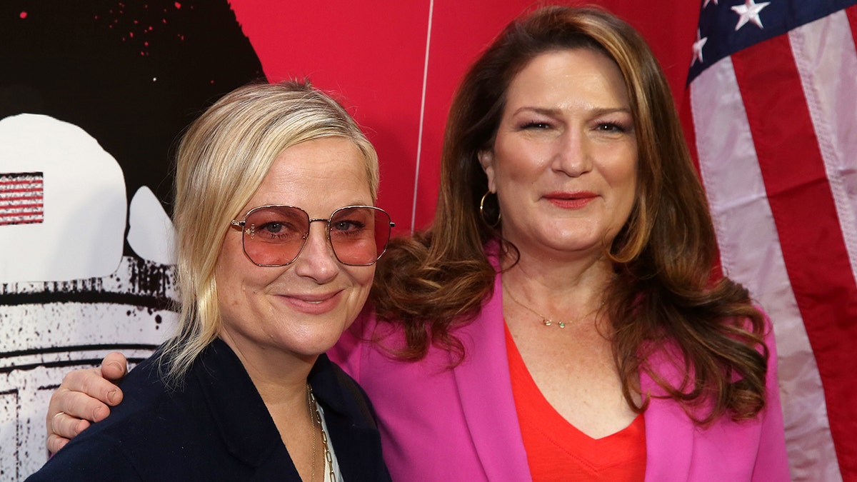 Amy Poehler and Ana Gasteyer posing together at a red-carpet style event, smiling in front of a patriotic backdrop with an American flag.