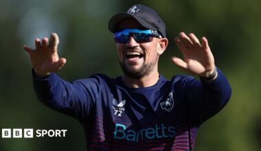 Harry Finch smiles with both arms raised in front of him, wearing a blue baseball cap, dark sunglasses and a blue cricket kit