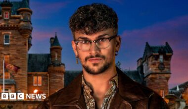 Jack Butler is looking at the camera. He has short brown curly hair and is wearing glasses, a patterned shirt and a brown leather jacket. He is standing in front of a castle.