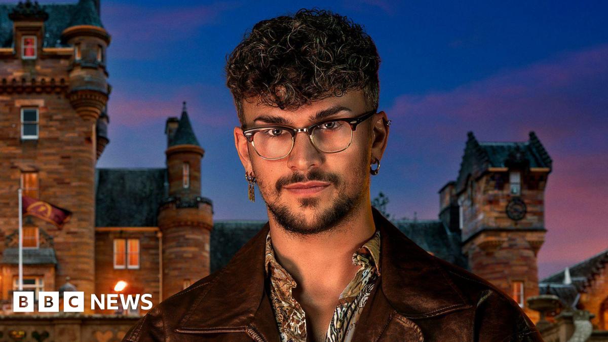 Jack Butler is looking at the camera. He has short brown curly hair and is wearing glasses, a patterned shirt and a brown leather jacket. He is standing in front of a castle.