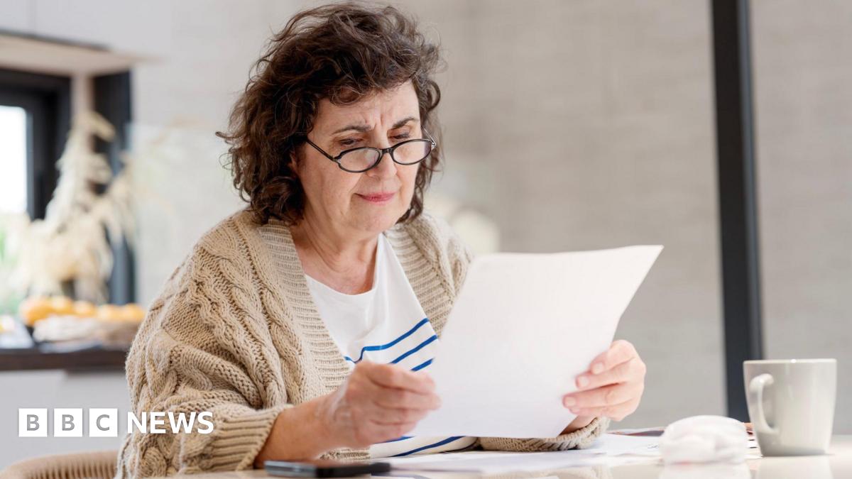 An older woman wearing glasses sits at a table looking at a piece of paper.