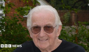 Ian Chatten smiles at the camera. He has short grey hair and wears sunglasses along with a black T-shirt. He is standing in front of a building with bushes and trees behind him.