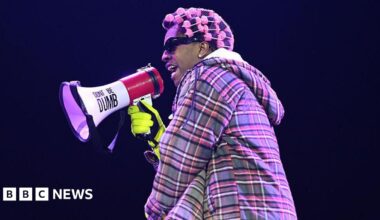 A$AP Rocky performing on stage. He wears a checked jacket and has pink rollers in his hair. He is holding a microphone with 'Don't Be Dumb' on it.