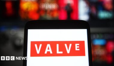 A phone with a white background and the orange logo of Valve Corporation, agianst a blurred background of games on the Steam platform.