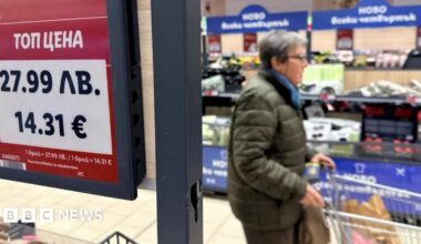 In a supermarket, a woman is blurry in the background, while in the foreground are two price tags showing both Lev and euros