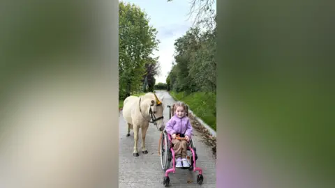 Ann Reel Hollie in her hot pink wheelchair, she is wearing a purple jacket and has her long brown hair tied in pigtails. She is holding the lead of her pony Rosco, who is a light coloured pony wearing a unicorn shaped hat. They are outside. 