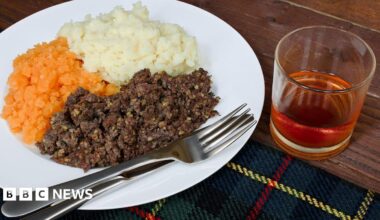 A white plate with a knife and fork, with brown haggis, mashed orange turnip and white mashed potatoes sitting on a tartan cloth on top of a table. There is a glass of whisky beside the plate.