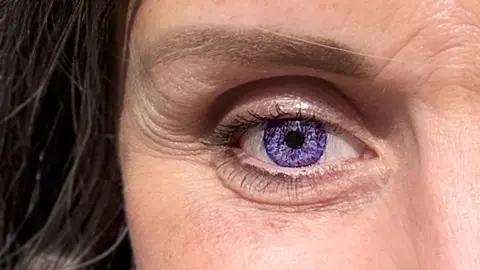 Charlie Flounders A close up of a woman's face. her eye has a purple prosthetic on it. The iris is a amethyst purple