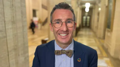 Muir is smiling. He wears glasses and has a navy jacket with a bow tie. He's wearing a colourful pin. He's in the hall in Stormont's parliament building. 