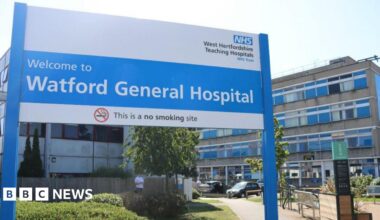 A large blue and white sign is in the foreground saying Welcome to Watford General Hospital" with the NHS logo above it. The hopsital - a concrete block of 4 storeys -is behind.