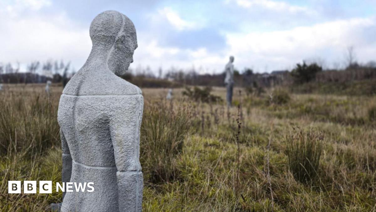 A rural landscape with grassy fields and shrubs under a cloudy blue sky. Scattered across the field are several pale, human-shaped sculptures, some standing upright and others partially submerged in the ground.