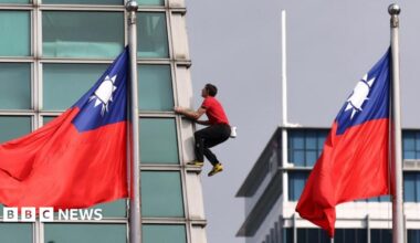 Alex Honnold stands on top of the Taipei 101 skyscraper building in a red t-shirt and dark trousers. There is only blue sky behind him.
