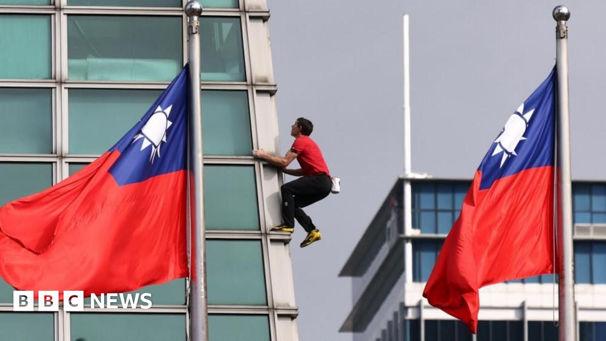 Alex Honnold stands on top of the Taipei 101 skyscraper building in a red t-shirt and dark trousers. There is only blue sky behind him.