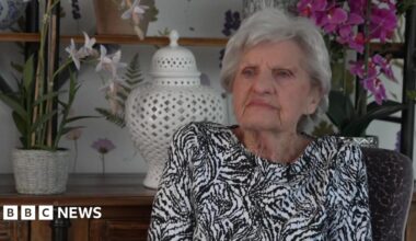 A woman with grey wears a black and white top and sits in front of an oak dresser with an orchid and vase on it.