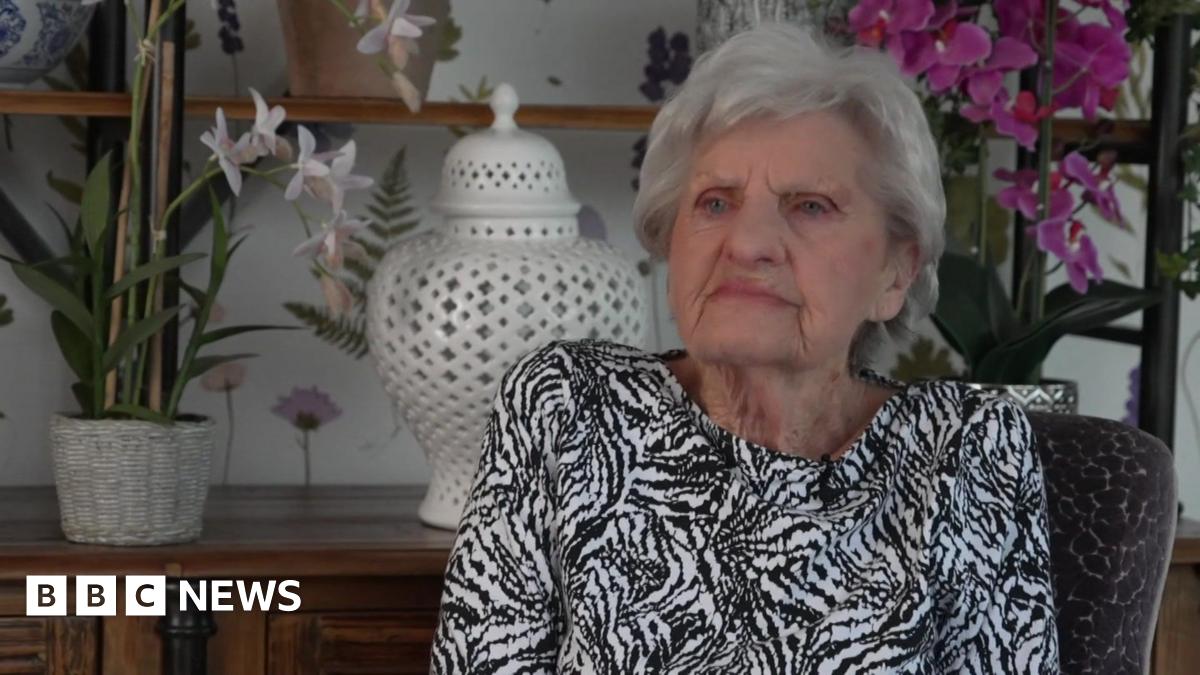 A woman with grey wears a black and white top and sits in front of an oak dresser with an orchid and vase on it.