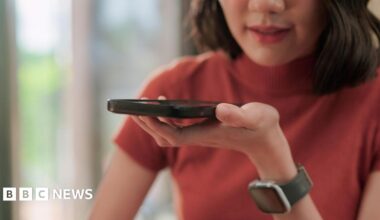 A woman wearing a red top speaks into the speaker of a smartphone she is holding in front of her.