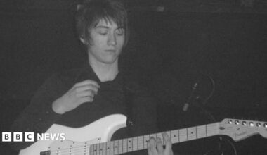 A young man with long dark hair playing a white guitar