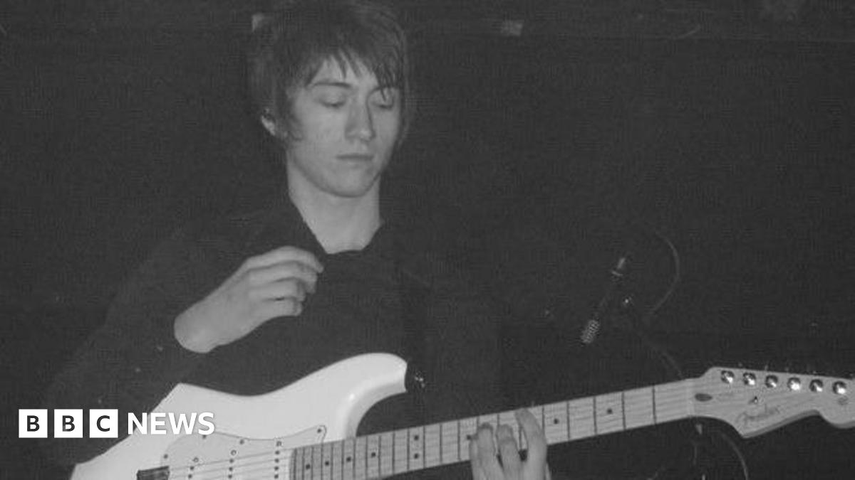 A young man with long dark hair playing a white guitar
