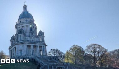 An ornate memorial building on a hill. Steps lead up to the domed building, while trees can be seen either side on a slope. The trail of an aeroplane cuts through a blue sky in the background.
