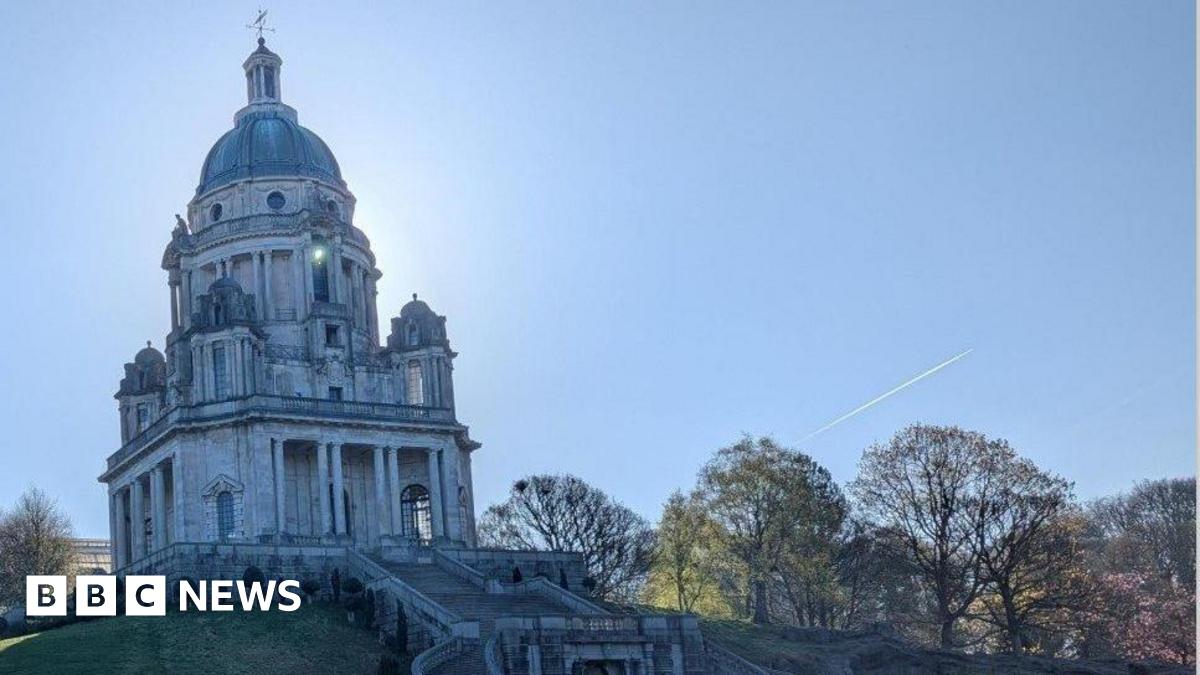 An ornate memorial building on a hill. Steps lead up to the domed building, while trees can be seen either side on a slope. The trail of an aeroplane cuts through a blue sky in the background.