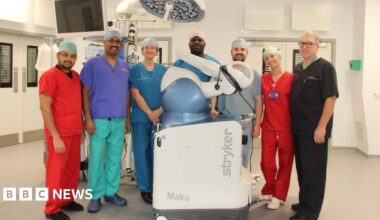Andrew Port is a man with short white hair and glasses and is wearing dark scrubs. Next to him there are four woman and two men wearing scrubs of different colours. They are stood in a surgical room with the white and blue Mako robotic arm.