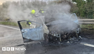 The picture shows a badly burned car on the side of a road. The front of the vehicle is almost completely destroyed, with the engine area exposed and charred. Thick smoke is rising from the wreckage, suggesting the fire has only recently been put out. The car doors are open, and the paintwork is scorched and peeling. Two firefighters wearing high-visibility helmets and protective gear are standing behind the car, partially obscured by the smoke.