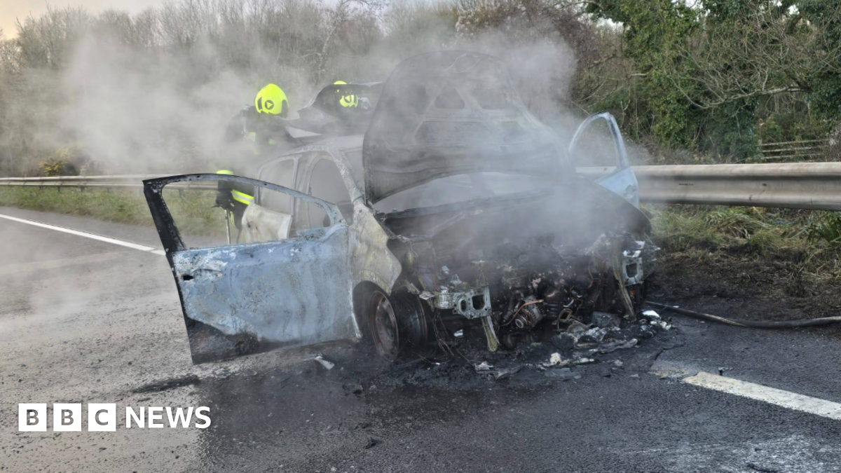 The picture shows a badly burned car on the side of a road. The front of the vehicle is almost completely destroyed, with the engine area exposed and charred. Thick smoke is rising from the wreckage, suggesting the fire has only recently been put out. The car doors are open, and the paintwork is scorched and peeling. Two firefighters wearing high-visibility helmets and protective gear are standing behind the car, partially obscured by the smoke.