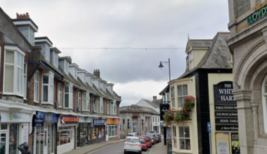 A general view looking down a street in a town centre. Shops line each side of the street. Cars are parked in on street car parking bays. A person is walking along a pavement.