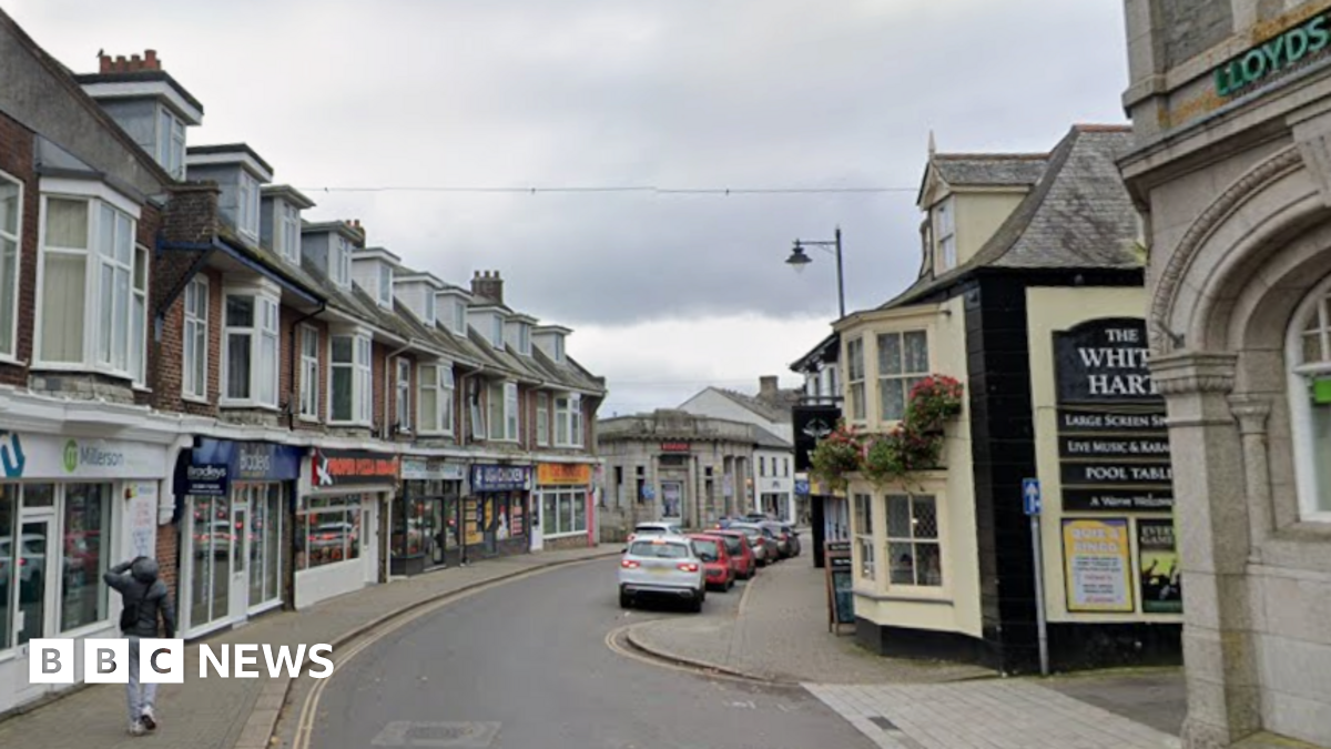 A general view looking down a street in a town centre. Shops line each side of the street. Cars are parked in on street car parking bays. A person is walking along a pavement.
