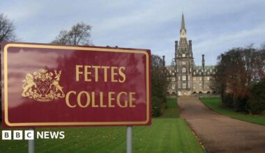 A red sign with "Fettes College" written in gold next to a coat of arms. The central turret of the stone school is visible at the end of a long pathway.