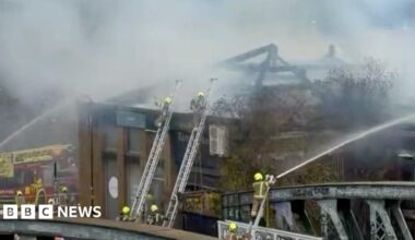 Firefighters on ladders wearing helmets spray water from hoses onto a building. A red fire engine is on the left